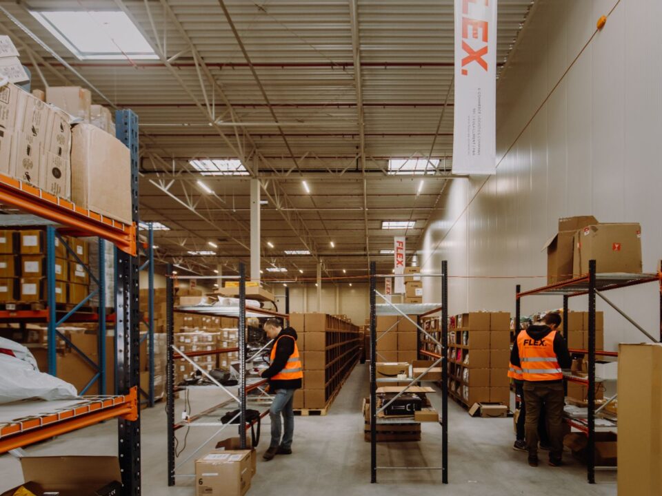 Workers in orange vests organize shelves in a spacious warehouse filled with boxes. The scene reflects orderliness and efficiency.